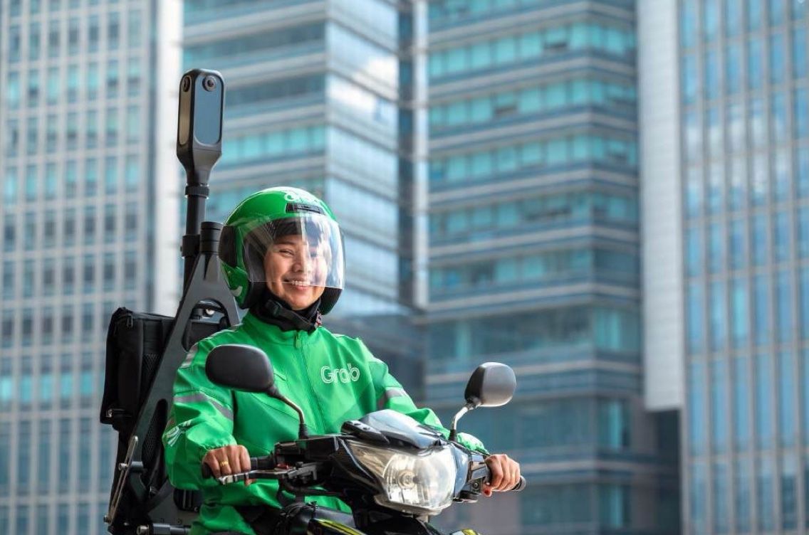 A smiling Grab rider on her scooter among glass towers, KartaCam mounted behind her