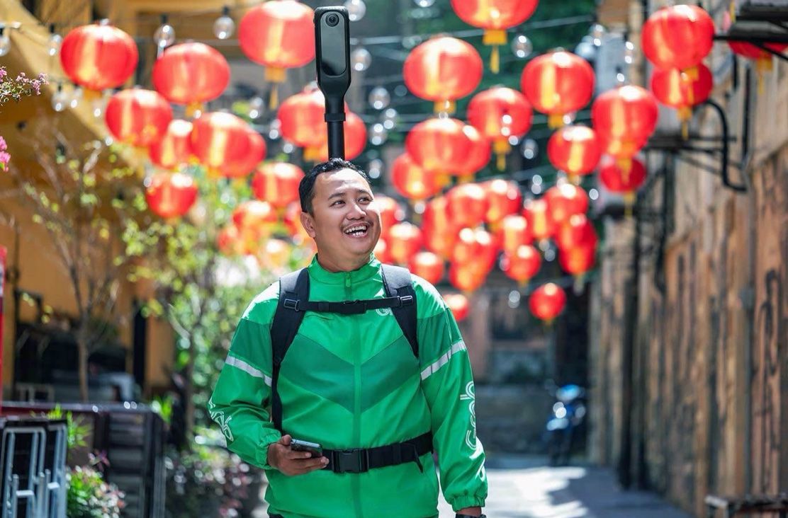 A Grab mapper in a green jacket smiles under red lanterns in a Kuala Lumpur alley, KartaCam 2 mounted above his backpack