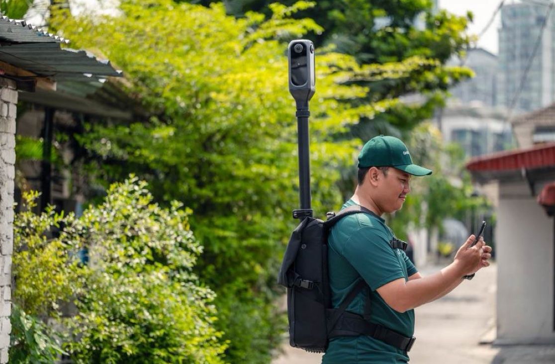 A mapper in a green cap checks his phone beside a leafy house