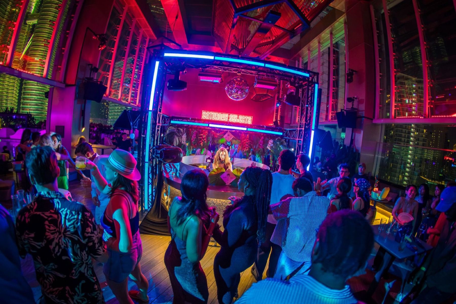 A crowd dancing in front of the Saturday Selects DJ booth under red and blue lights